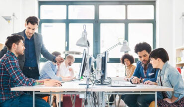 A set of employees are discussing about business process sitting around the round table, table lamps, computers, smart phones can be seen.