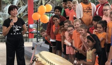 Some students are in a music class outside the classroom. 1 girl is playing the drum. Other students are watching her. The teacher is standing with a mike nearby. All the students belong to mixed ethnicity.