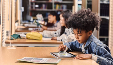 A small boy, along with some other students, is learning on the tablet, sitting on the table.