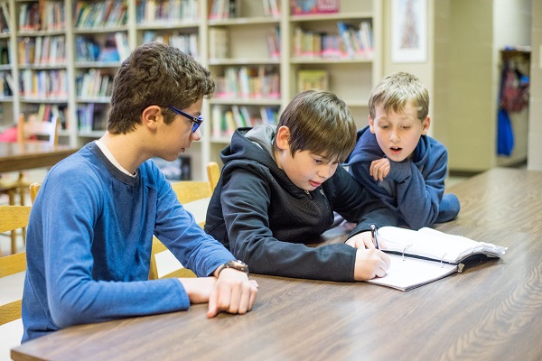 Three students sitting on the table and watching a book which is kept by a boy sitting in the middle.